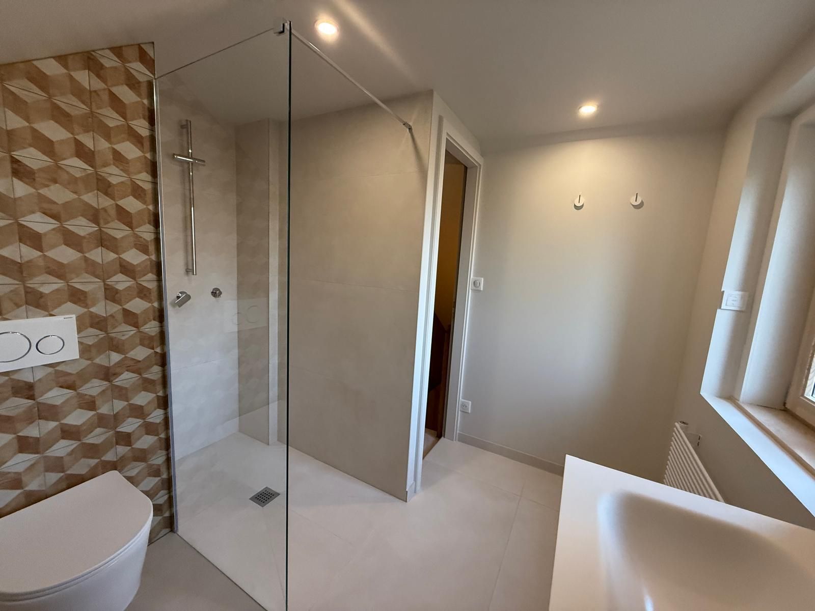 Salle de bains moderne avec douche en verre, WC suspendu, carrelage beige et grande fenêtre donnant sur un coin lavabo lumineux