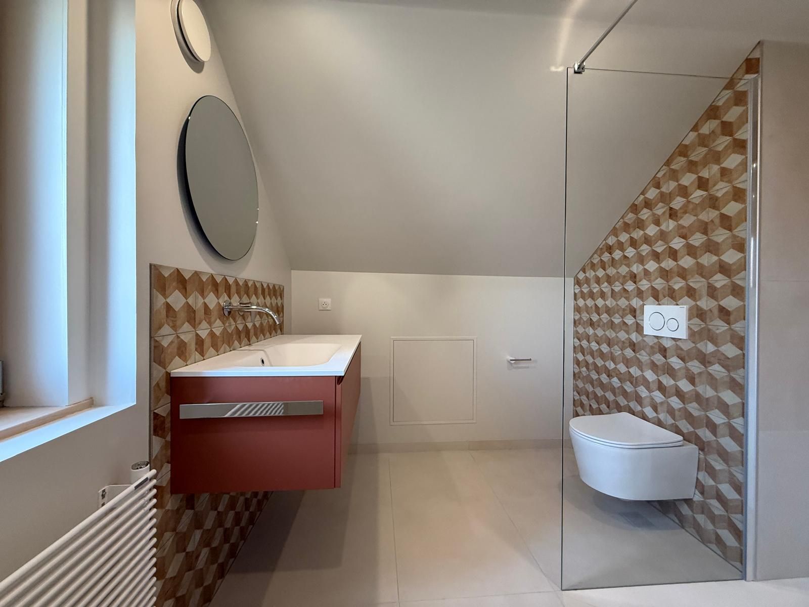 Salle de bains moderne avec lavabo suspendu, miroir rond, toilettes murales et carrelage beige à motifs.