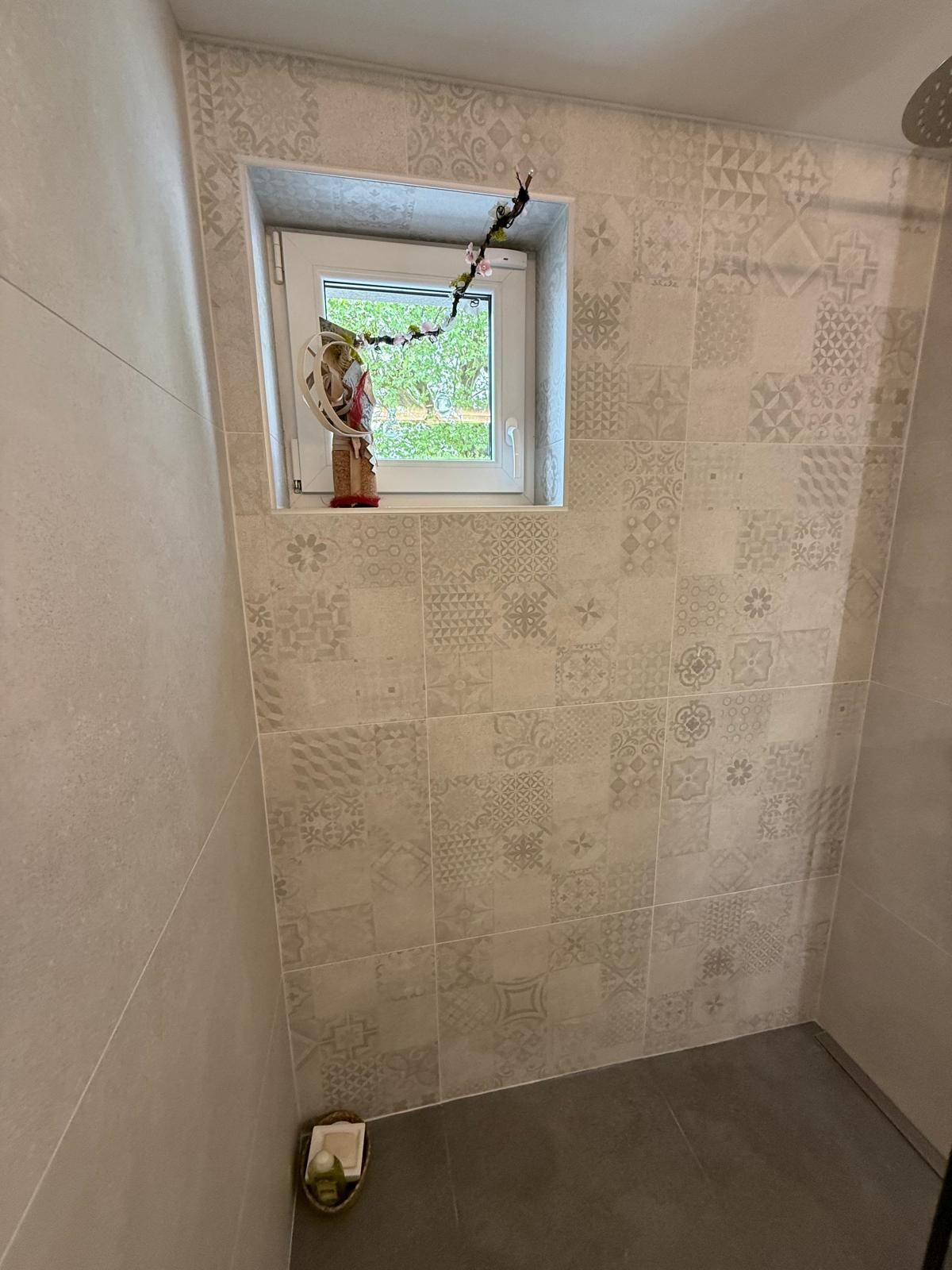 Petite salle de bains carrelée avec fenêtre, porte-papier toilette et sol foncé.
