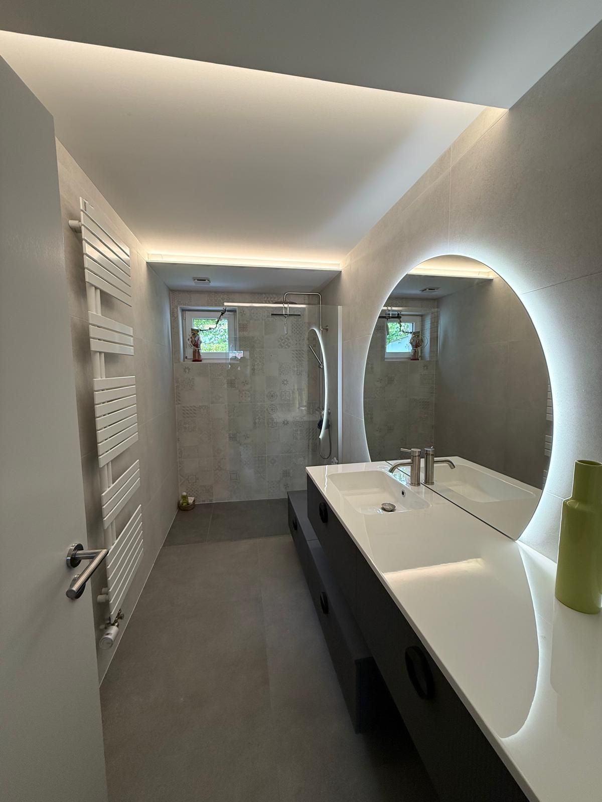 Salle de bains moderne avec meuble-lavabo blanc, miroir rond, douche et petites fenêtres baignées d'une lumière douce.