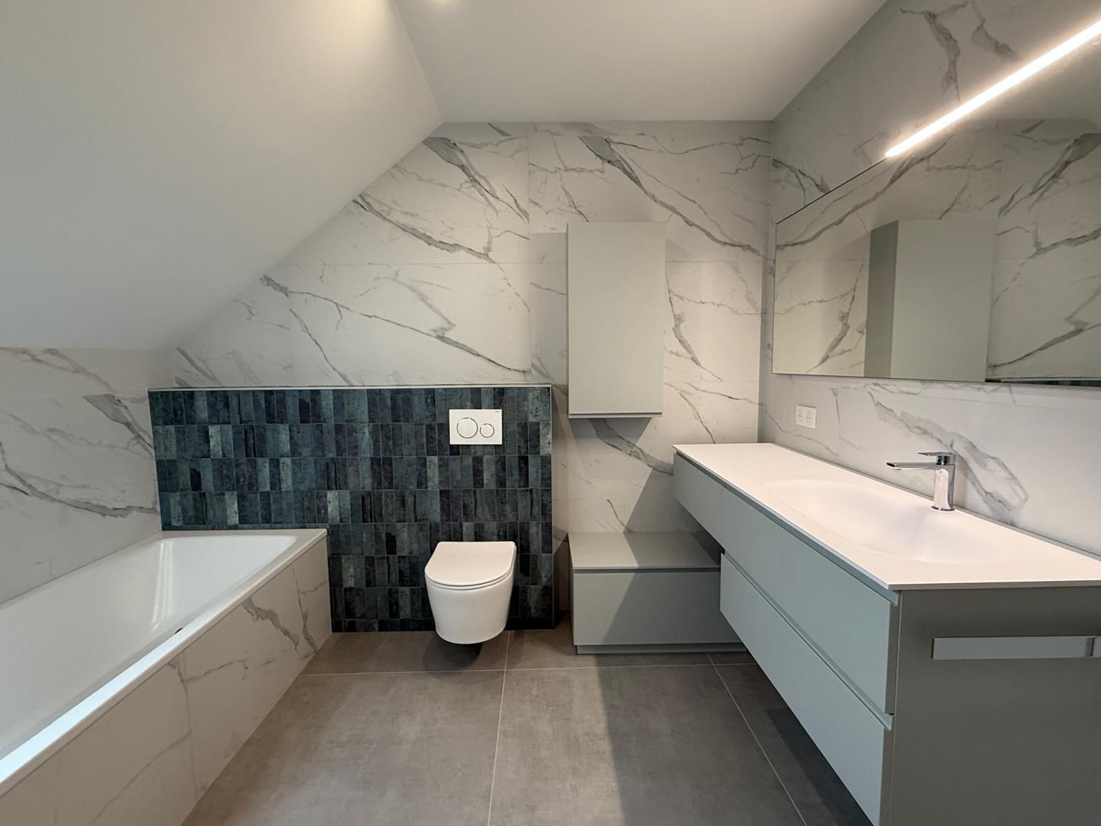 Salle de bains moderne avec murs en marbre, baignoire, toilettes et double vasque sous un plafond mansardé
