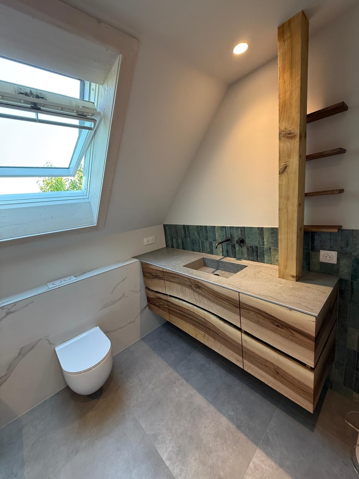 Petite salle de bains avec plafond mansardé, puits de lumière, toilettes et meuble-lavabo en bois avec étagères et miroir.