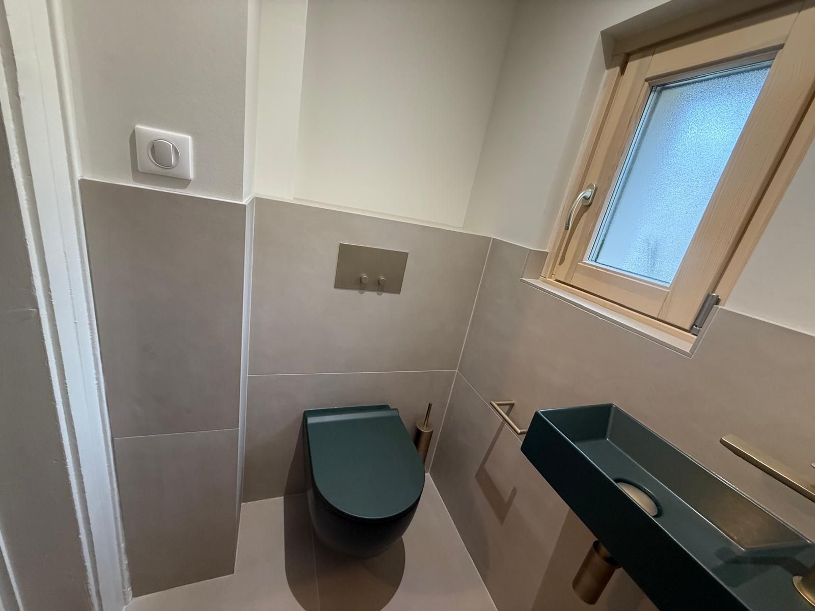 Petite salle de bains carrelée avec toilettes suspendues, lavabo et fenêtre dépolie.