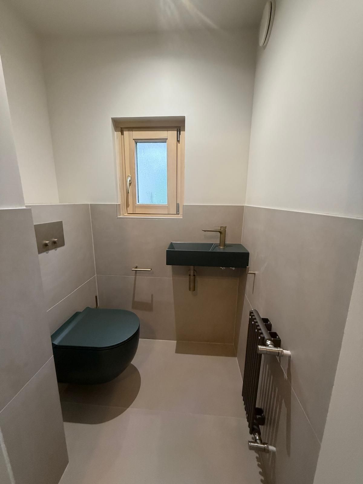 Salle de bains étroite avec toilettes, lavabo, fenêtre et radiateur, murs blancs et beiges.