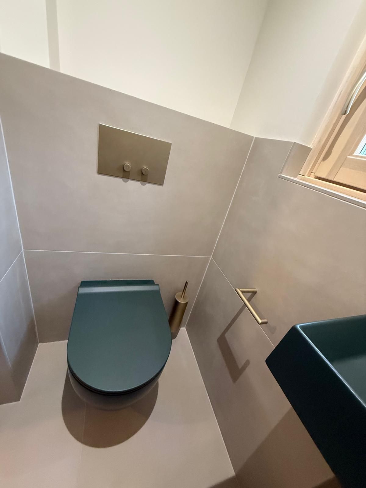 Toilettes modernes avec siège turquoise, murs carrelés gris et plaque de chasse d'eau en métal sous une petite fenêtre