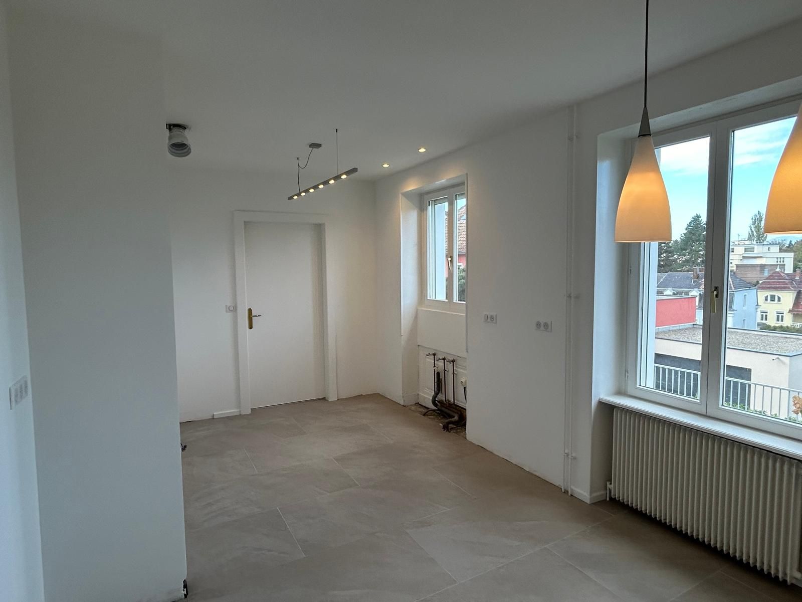 Pièce blanche vide avec de grandes fenêtres, un radiateur et une suspension lumineuse.