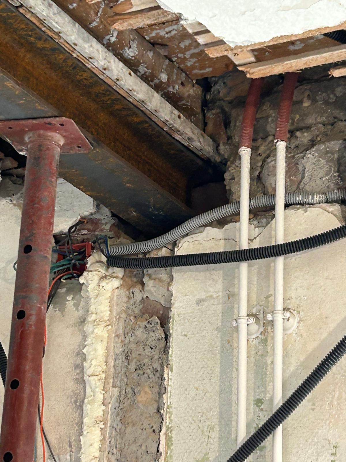 Poutres et tuyaux apparents au plafond dans un intérieur inachevé, avec des câbles courant le long d'un mur en béton.