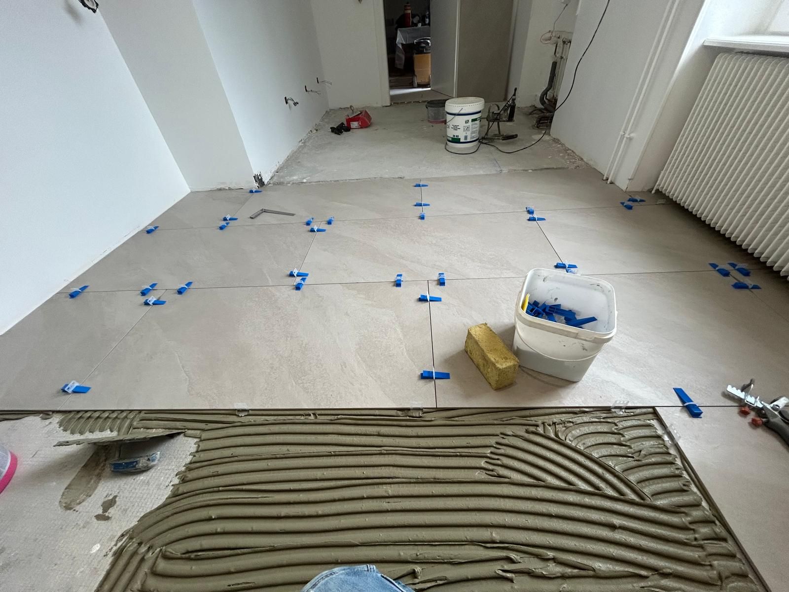 Sol carrelé en cours de rénovation : colle appliquée, croisillons bleus et seaux de peinture dans un couloir