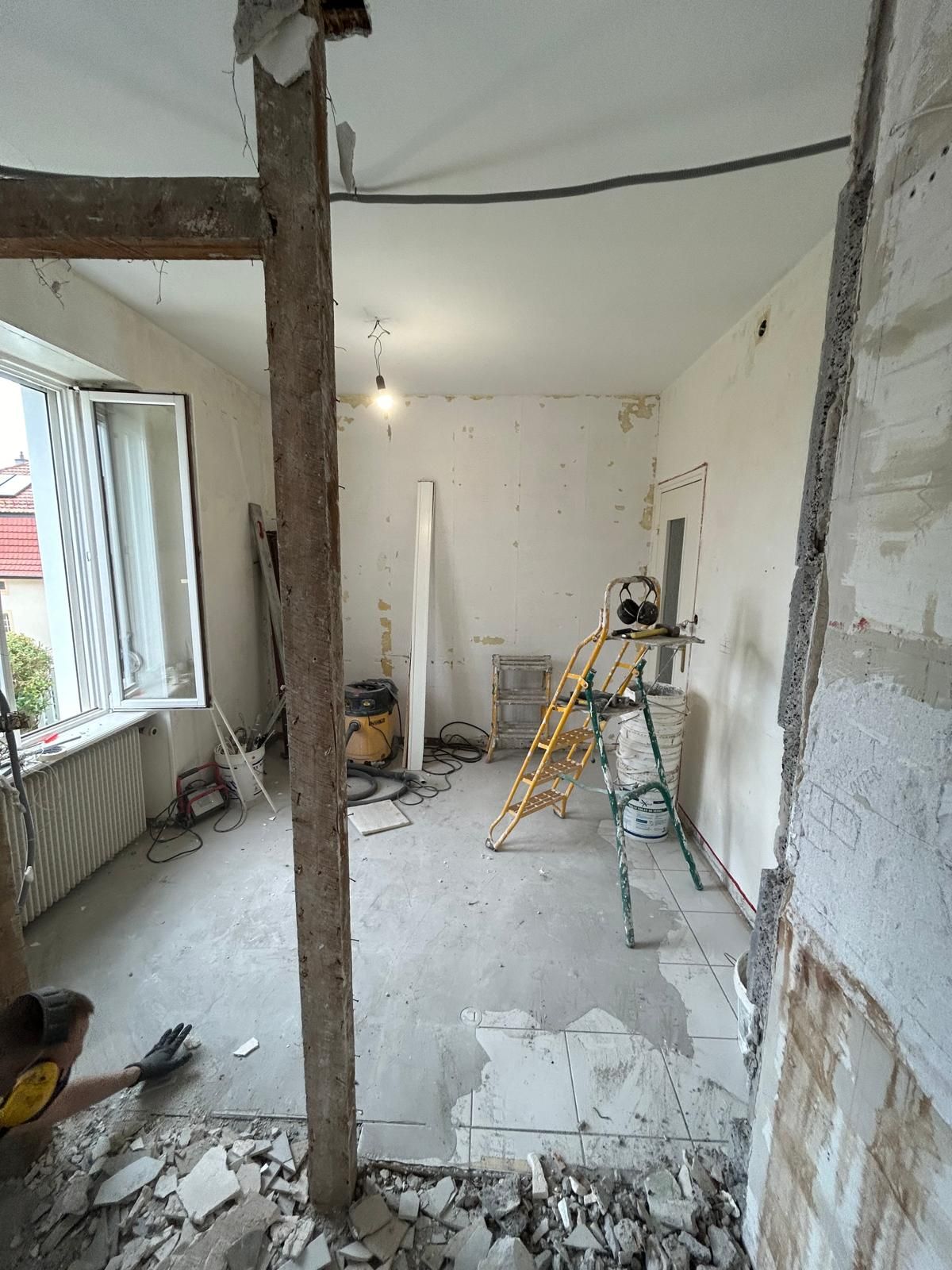 Travaux de rénovation en cours dans une pièce étroite aux murs apparents, avec des débris, une échelle et des outils près d'une fenêtre.