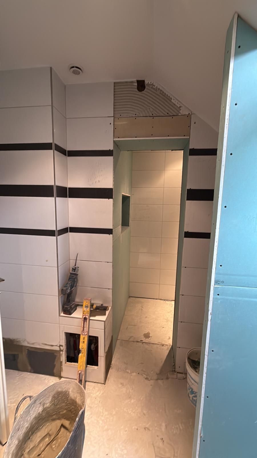Salle de bains carrelée avec une porte étroite, des murs blancs à rayures et un espace lavabo et toilettes visible.