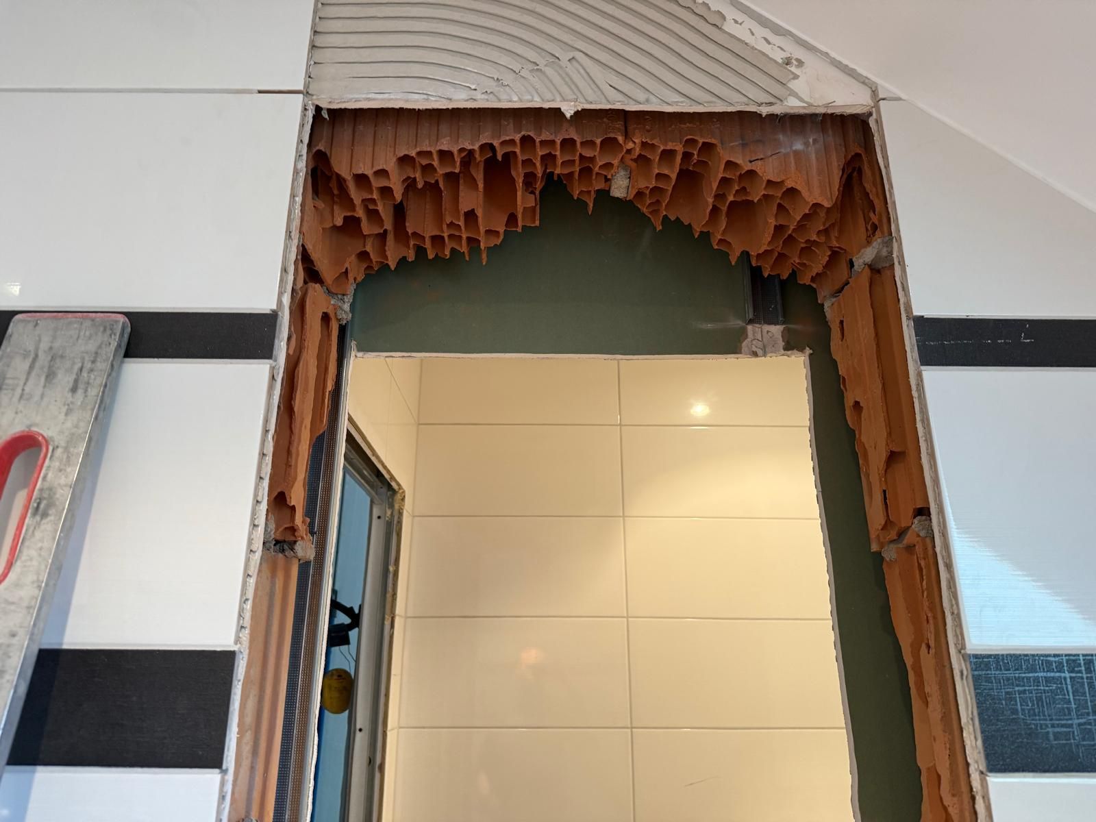 Porte endommagée avec briques cassées apparentes et débris autour du cadre dans un couloir intérieur carrelé