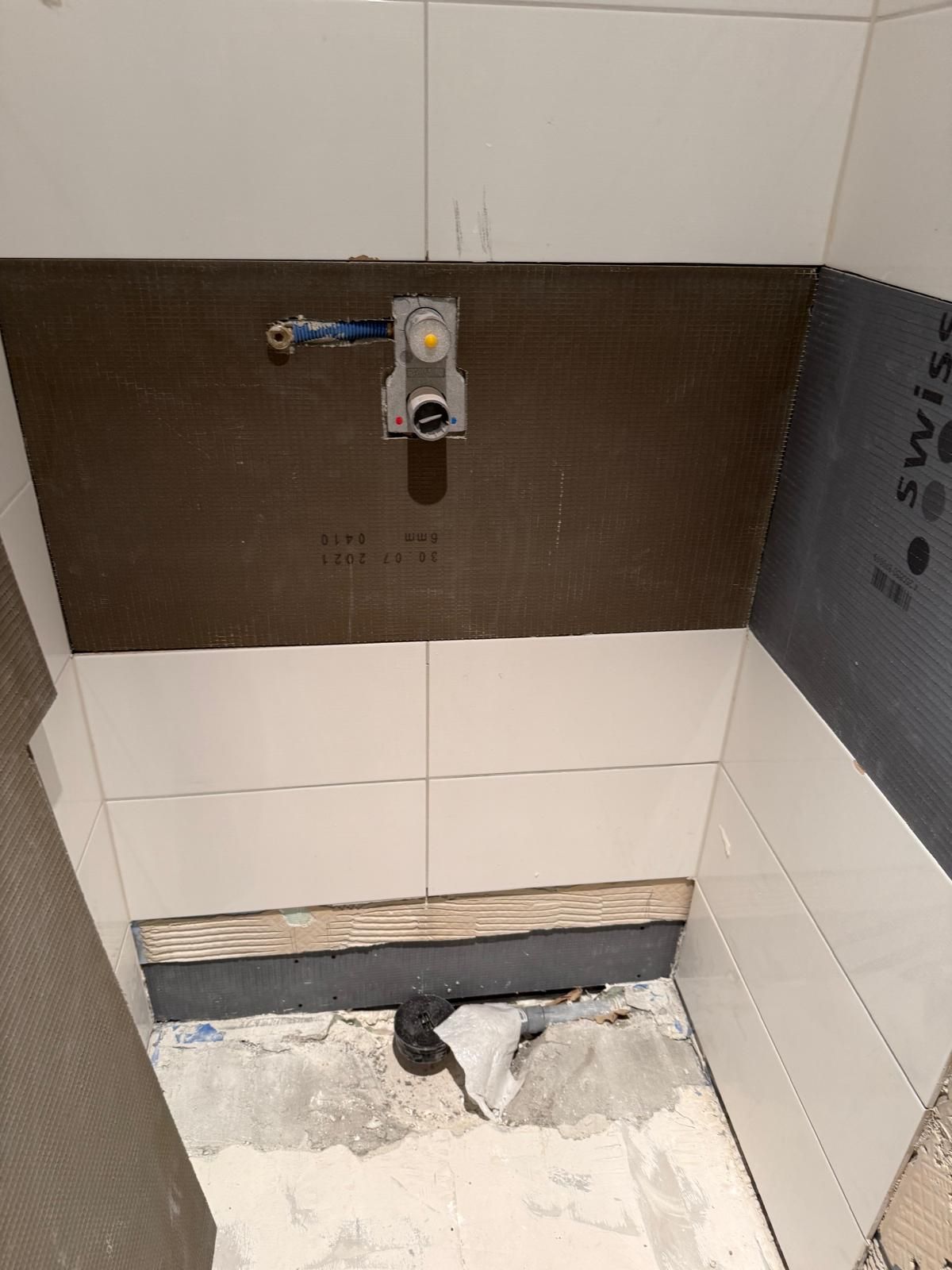 Mur de salle de bain partiellement carrelé avec plomberie apparente et ouverture dans le sol non fini.