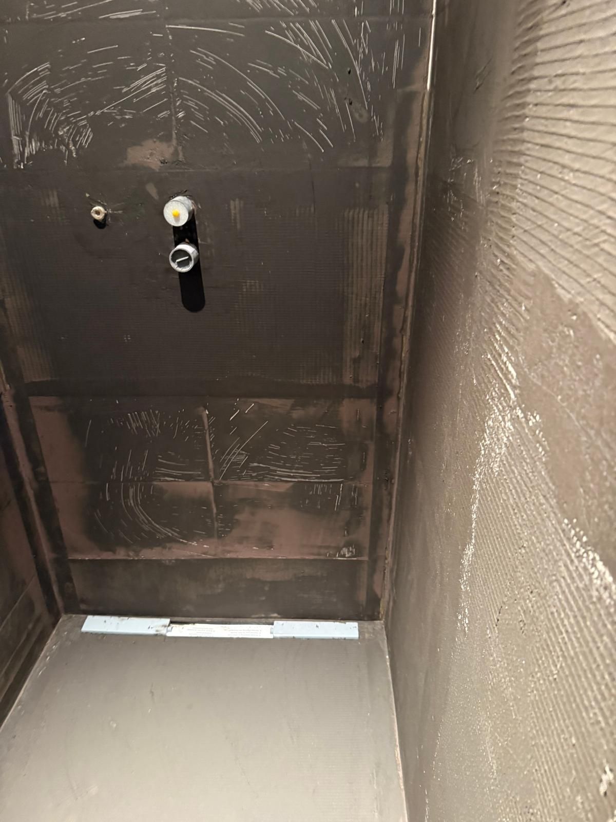 Cabine de douche vide avec murs carrelés foncés, robinet mural et panneau de sol clair.