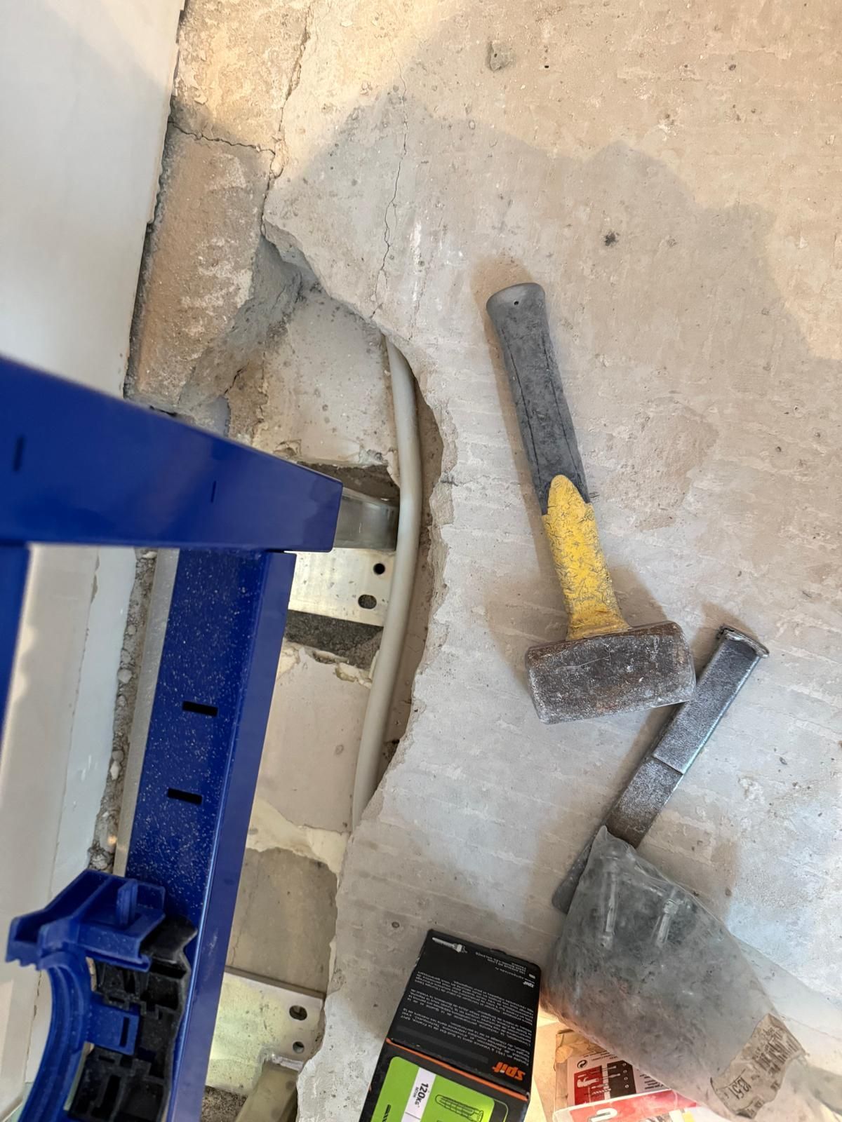 Marteau et outils à côté d'une échelle bleue sur un sol en béton, avec un sac en plastique et de petits objets à proximité.