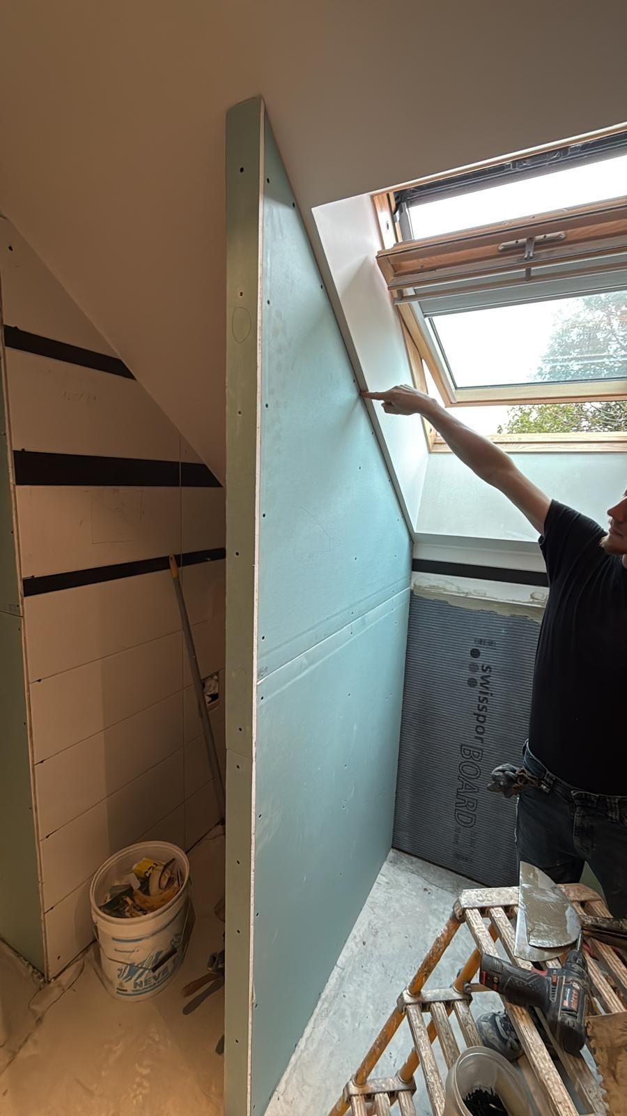 Une personne installe un grand panneau bleu-vert à côté d'une lucarne ouverte dans une cage d'escalier étroite.