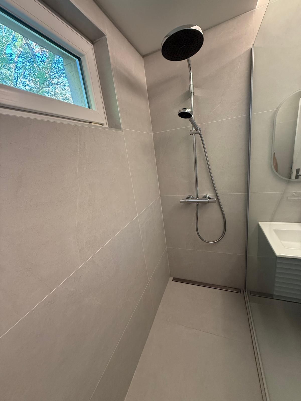 Petite salle de bain carrelée avec douchette murale et petite fenêtre.