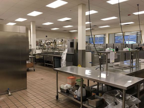 Empty commercial kitchen with stainless steel counters, sinks, and equipment under bright fluorescent lights.