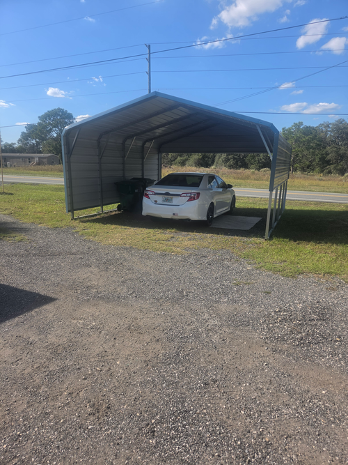 White car parked under a metal carport on a gravel lot under a blue sky.
