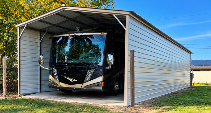 Motorhome parked inside a metal carport beside a grassy field under a blue sky