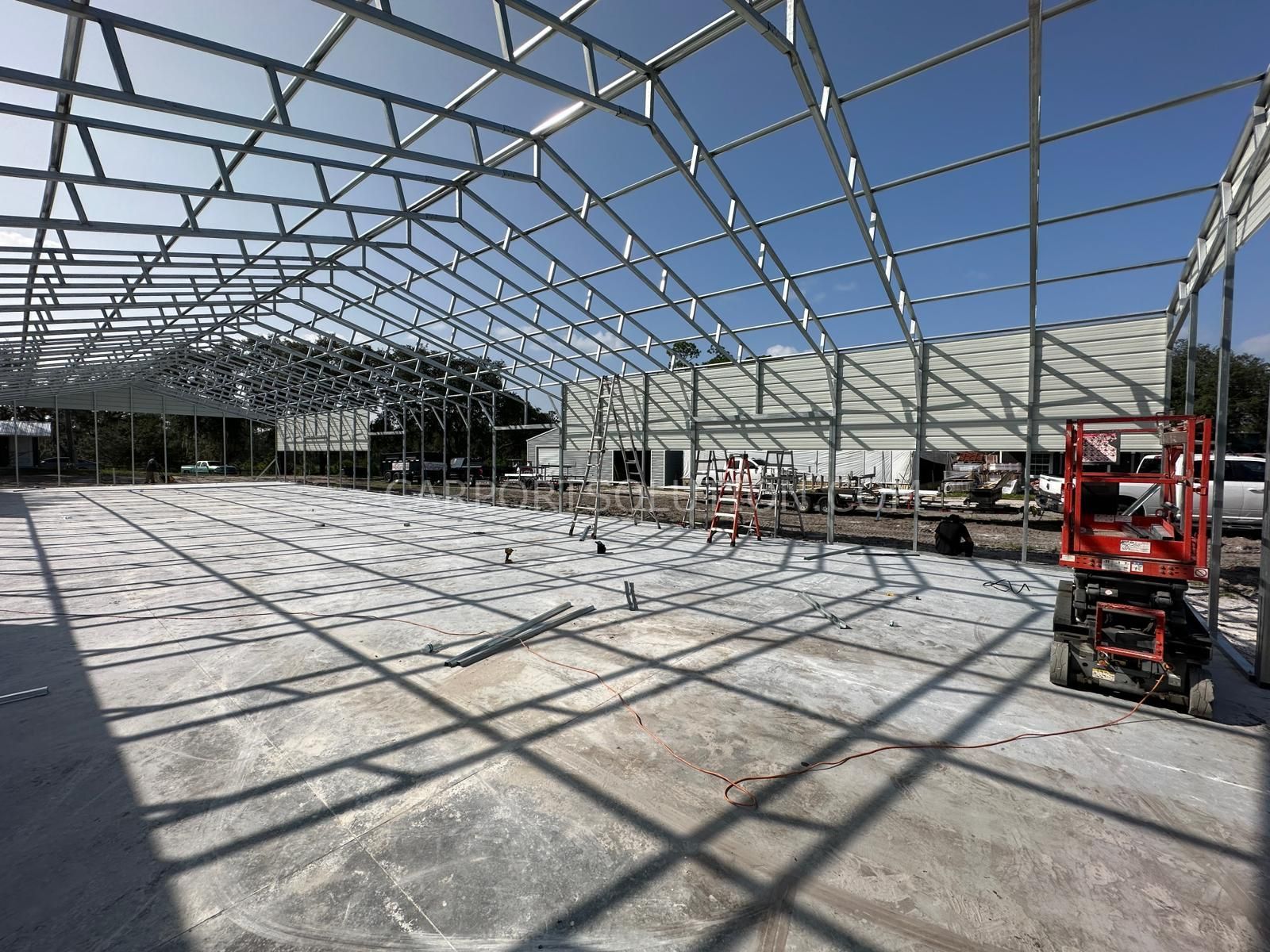 Large steel frame structure under construction on a concrete slab, with a lift on the right and open sky above.