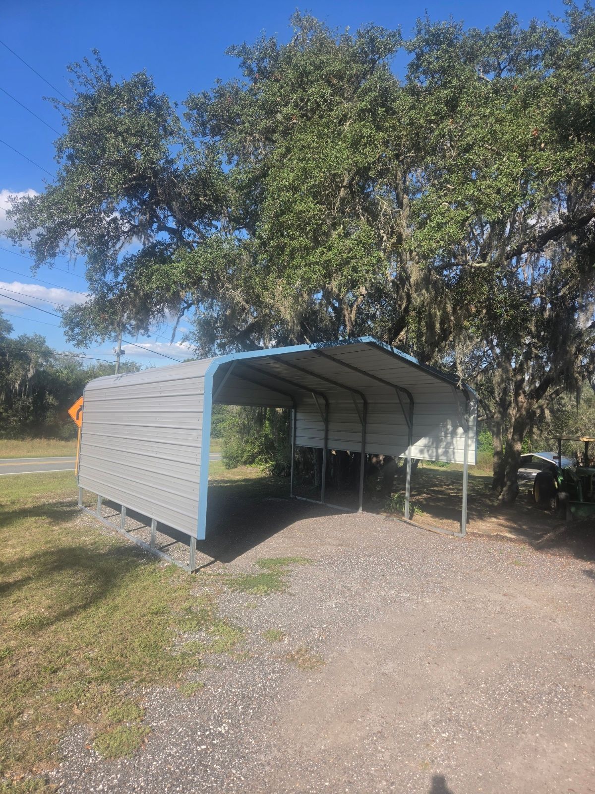 Open-sided metal carport on a gravel drive, surrounded by trees under a clear blue sky