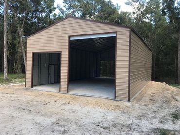 Open-sided metal workshop shed with tan siding, sitting on a dirt lot near trees