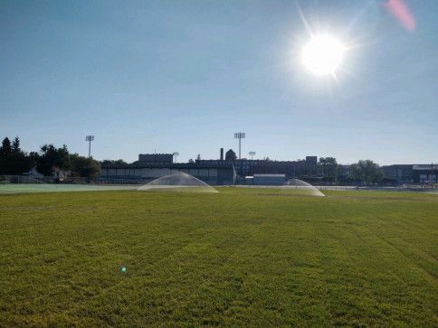 Large sprinklers water a bright green athletic field under a clear, sunny sky, with stadium lights visible in the distance.