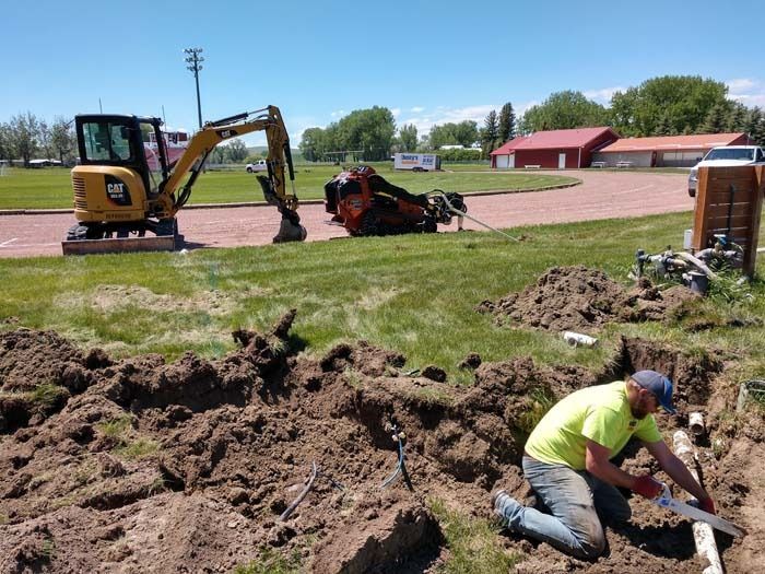 A worker in a high-visibility shirt kneeling in a trench, cutting a pipe, with heavy construction machinery parked nearby.