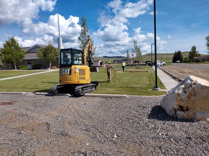 A yellow Caterpillar excavator sits on a grass lawn near a gravel lot, with two workers standing nearby on a sunny day.
