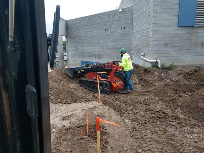 A worker in a high-visibility yellow jacket operates a compact orange tracked loader on a muddy construction site.