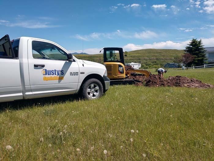 A white Justy’s construction truck parked in a grassy field next to an excavator digging a trench.