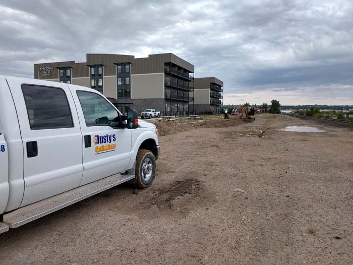 A white work truck parked on a dirt lot in front of modern, multi-story buildings under a cloudy sky.
