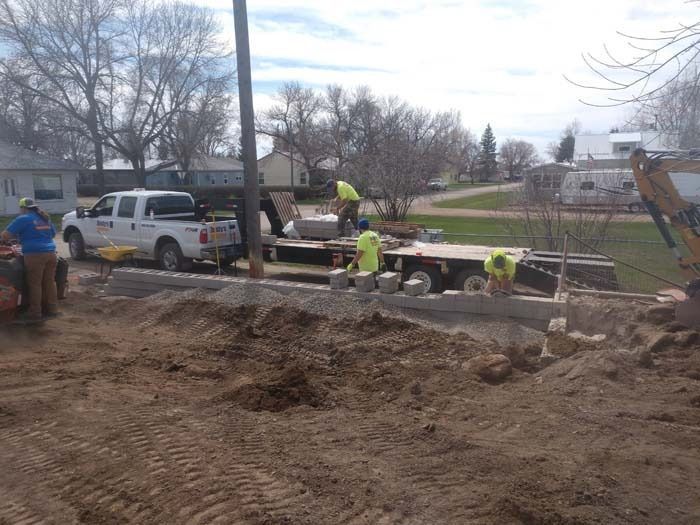 Three construction workers in neon vests install a block wall at a residential site with a truck and equipment nearby.