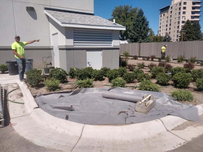 Two workers in neon shirts prepare a circular landscaping area with weed barrier fabric outside a building.