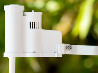 A white, plastic outdoor weather sensor mounted on a pole against a blurry, out-of-focus green garden background.