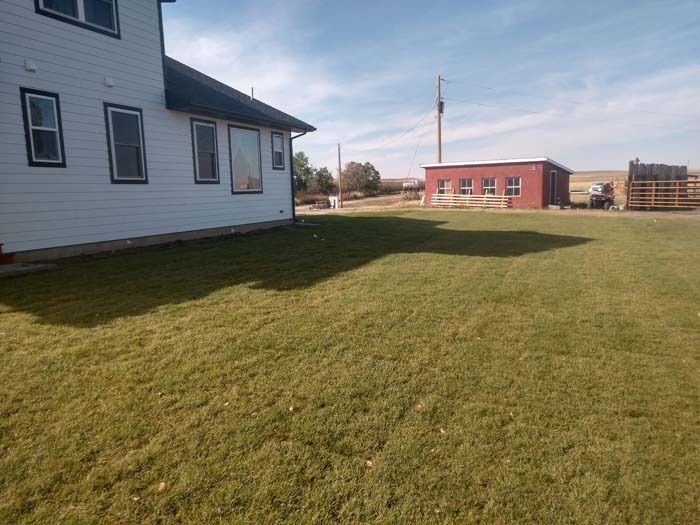 A white house with dark trim sits on a grassy yard, with a red barn in the distance under a blue sky.