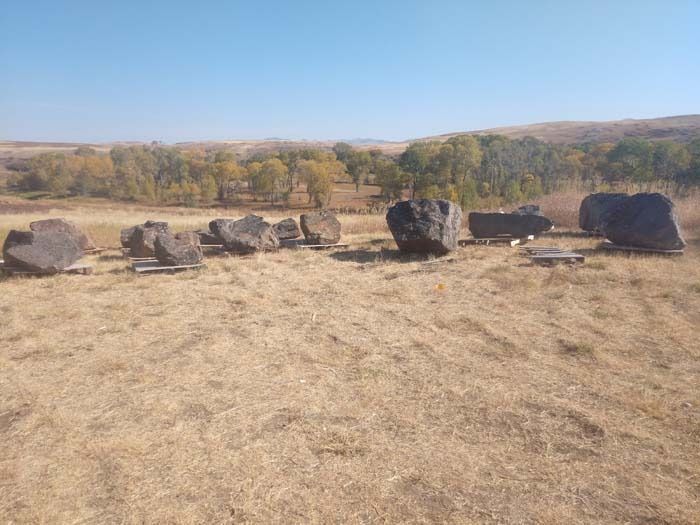 A row of large, dark boulders sits in a dry, grassy field against a backdrop of trees and hills under a clear blue sky.