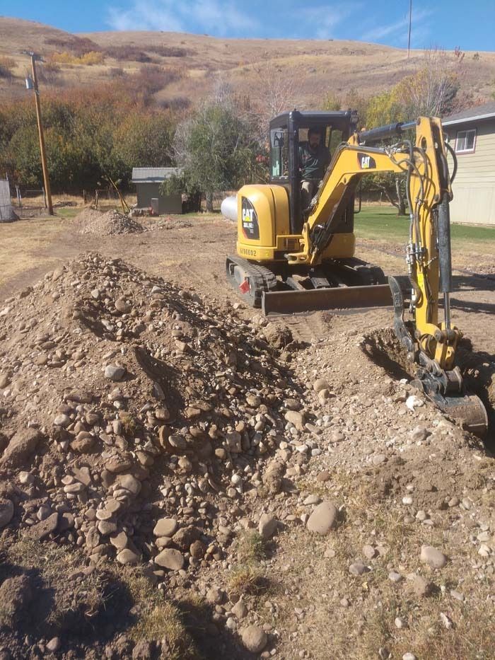 A yellow Caterpillar mini excavator sits on a dirt lot next to a mound of gravel, with hills in the background.