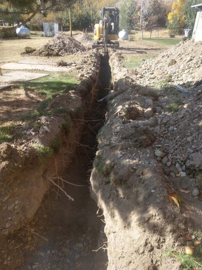 A yellow excavator digs a long, deep trench through a grassy yard toward the background.