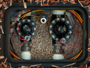 An in-ground irrigation valve box containing two black solenoid valves connected by pipes, surrounded by gravel and wiring.