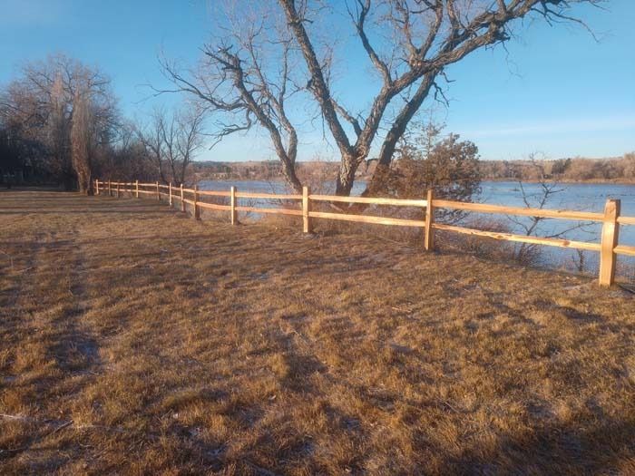 A two-rail wooden fence lines a grassy field overlooking a body of water under a clear, bright blue sky.