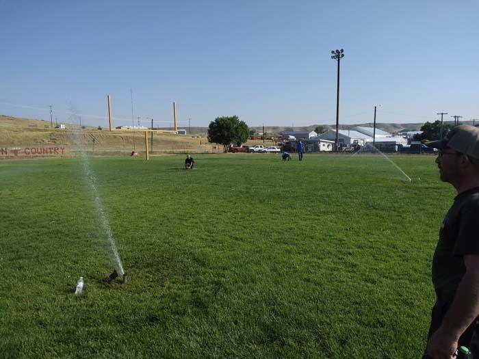 A person watches sprinklers watering a grassy field on a sunny day at a sports complex.