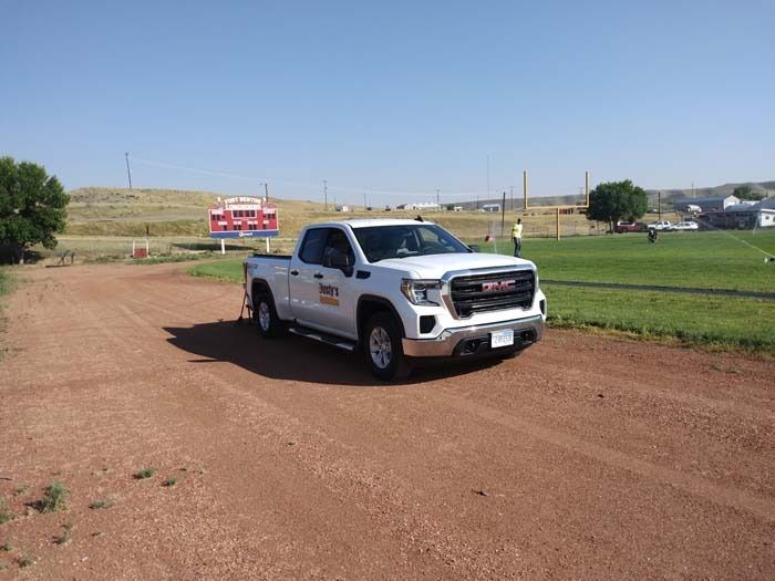 A white GMC pickup truck drives across a reddish dirt field next to a grass sports field and a scoreboard.