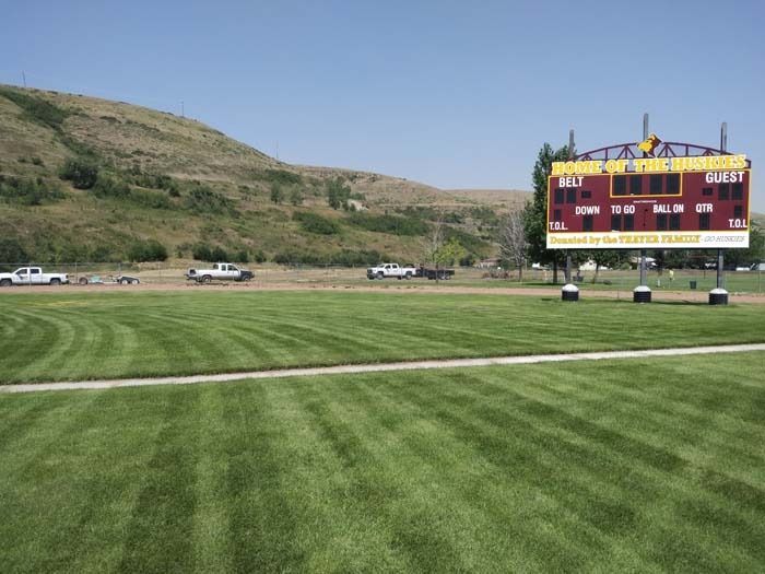 A football scoreboard stands next to a neatly mowed green field with a grassy hill and parked trucks in the background.