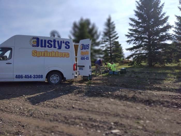A white Dusty's Sprinklers work van parked on a dirt lot with two employees working in the background near evergreen trees.