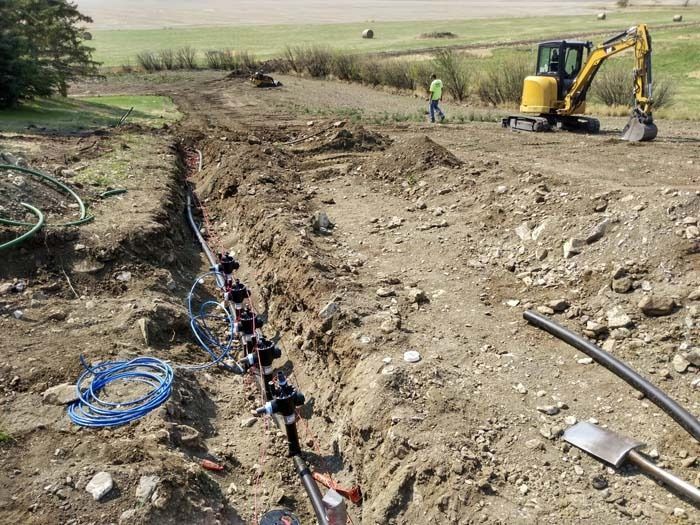 A yellow excavator operates in a rural field next to an open trench containing a newly installed irrigation valve system.