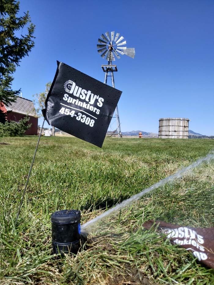 A sprinkler head sprays water in a grassy field, with a branded Justy's Sprinklers flag and a windmill in the background.