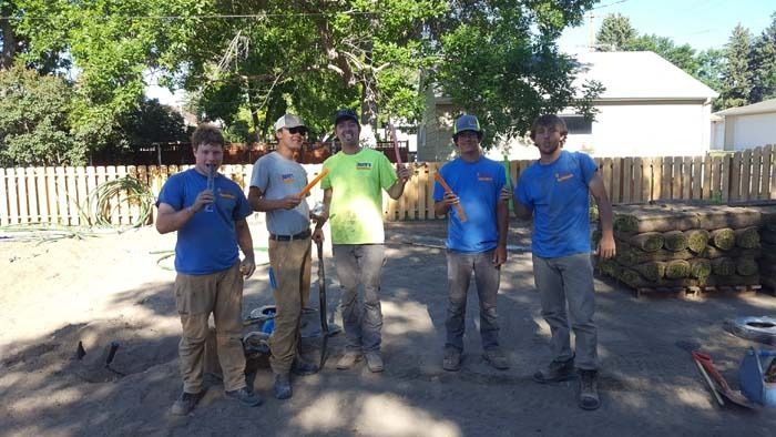 Five people in work clothes stand in a sunny, fenced-in construction area, posing with shovels and tools.