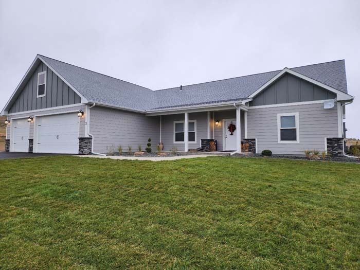 A modern, light gray single-story house with a white garage, dark stone accents, and a wide front porch on a grassy lot.