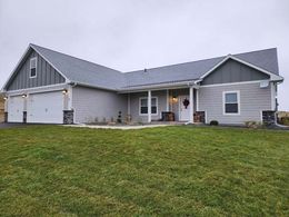 A light gray, single-story ranch house with a two-car garage, dark metal roof, and stone accents under a cloudy sky.